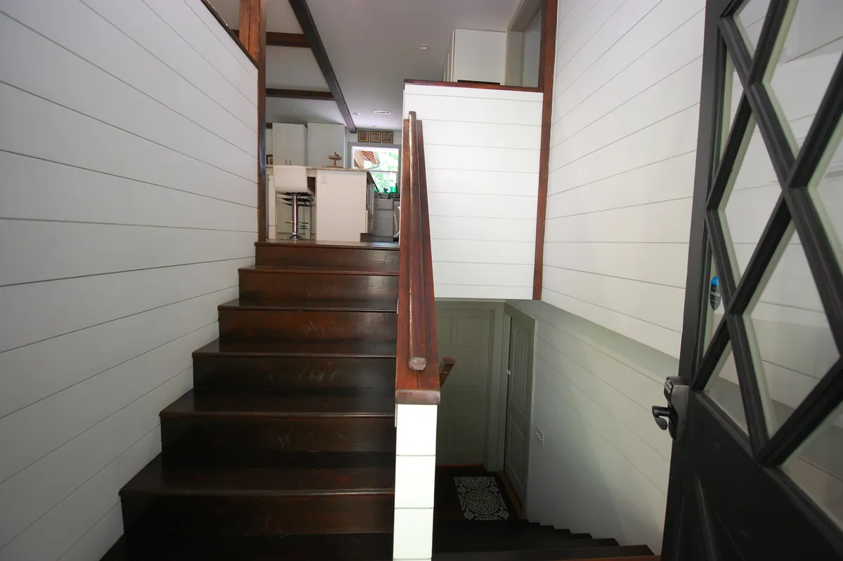 This image features a welcoming interior view centered on a staircase with a wood railing, light walls, and an open sightline to the kitchen. The space feels inviting and transitions naturally toward the outdoors, suggesting movement and activity.