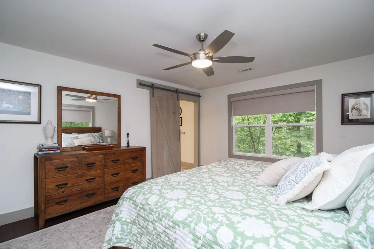 A bedroom featuring a bed with a green quilt, a wooden dresser, and a large window that frames natural views of the surrounding woodland. The room is filled with natural light, enhancing its tranquil, nature-connected atmosphere.