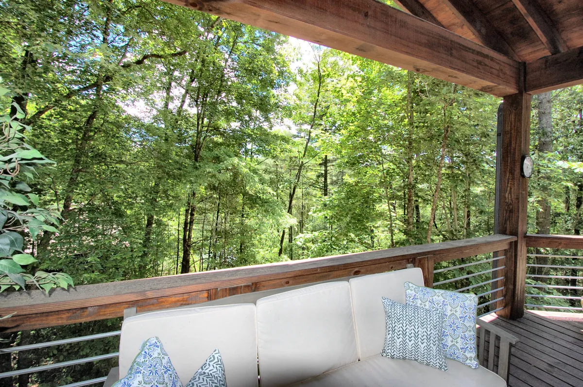 A serene wooden deck with a comfortable couch adorned with pillows, surrounded by greenery. The space is ideal for relaxing, reading, or simply soaking in the natural beauty and fresh mountain air.
