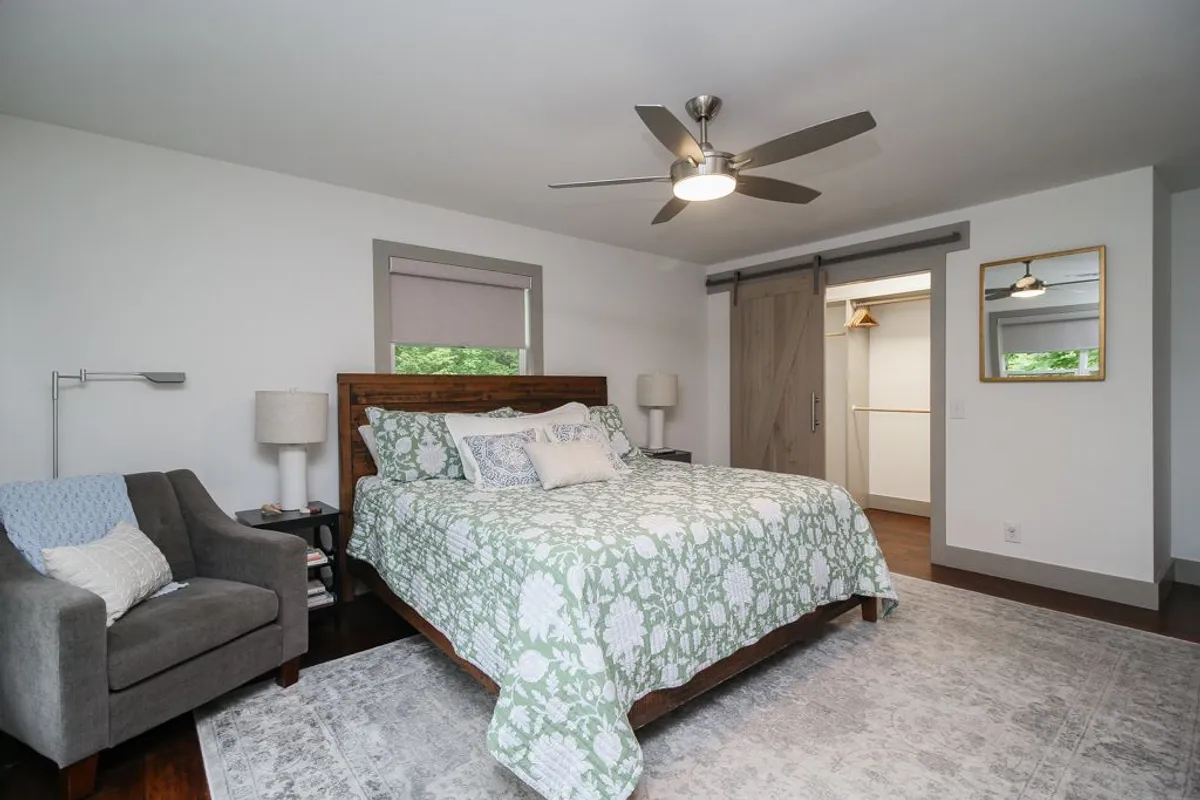 A cozy bedroom with a bed dressed in floral pattern bedding, armchair beside a window, soft lighting, and a sliding door leading to a closet. The ambiance is peaceful and tranquil, suggesting a restful retreat ideal for relaxation and privacy.