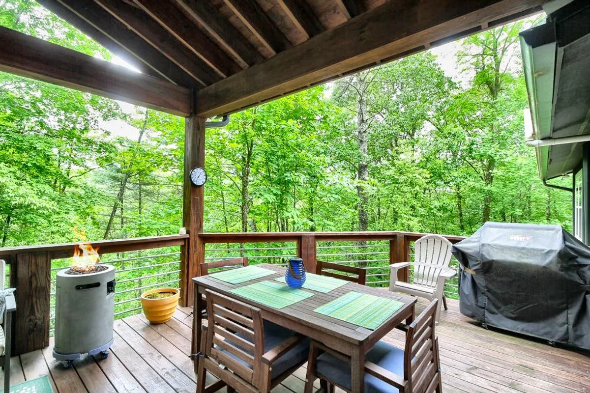 This image features an outdoor dining table, a fire pit, a gas grill, and is surrounded by lush greenery. The setting is inviting and peaceful, perfectly suited for outdoor dining, relaxing, and entertaining in a natural environment.