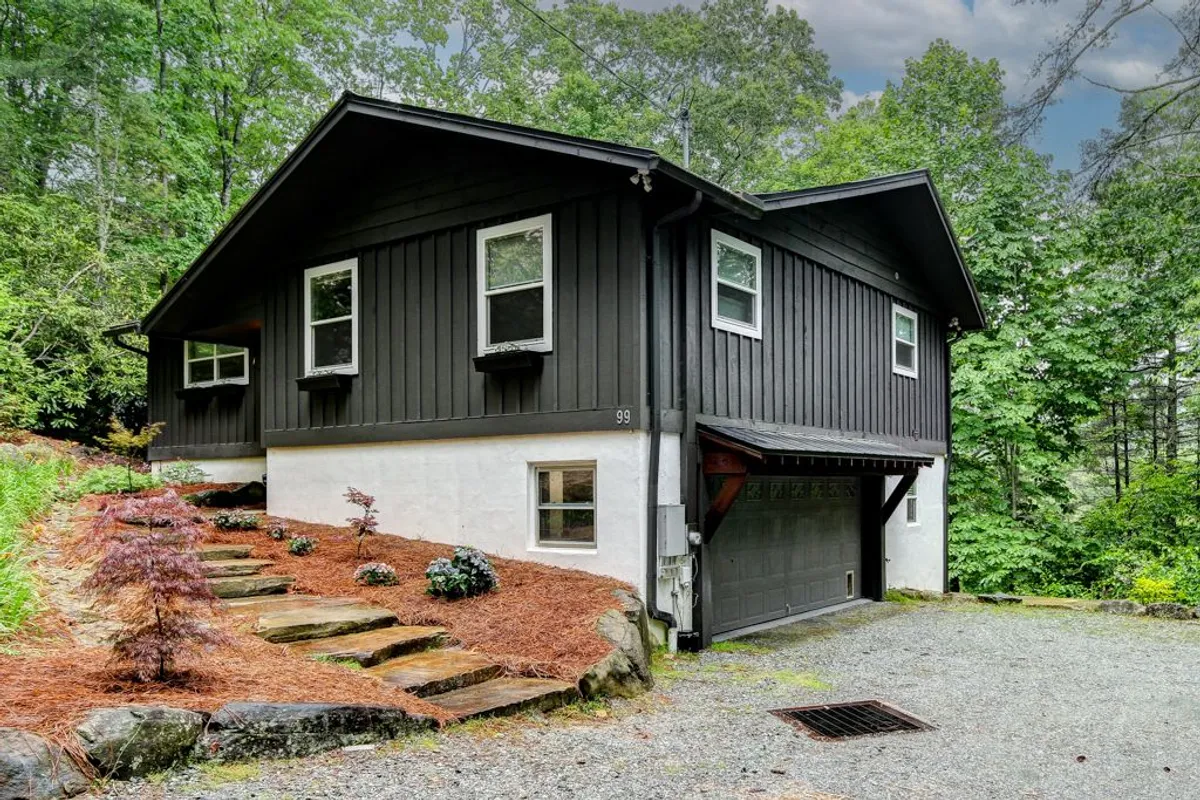 This exterior shot showcases the home’s black wooden siding, white stucco foundation, rocky steps, and lush green foliage surrounding the property. The gravel driveway invites guests into a serene and welcoming mountain setting.