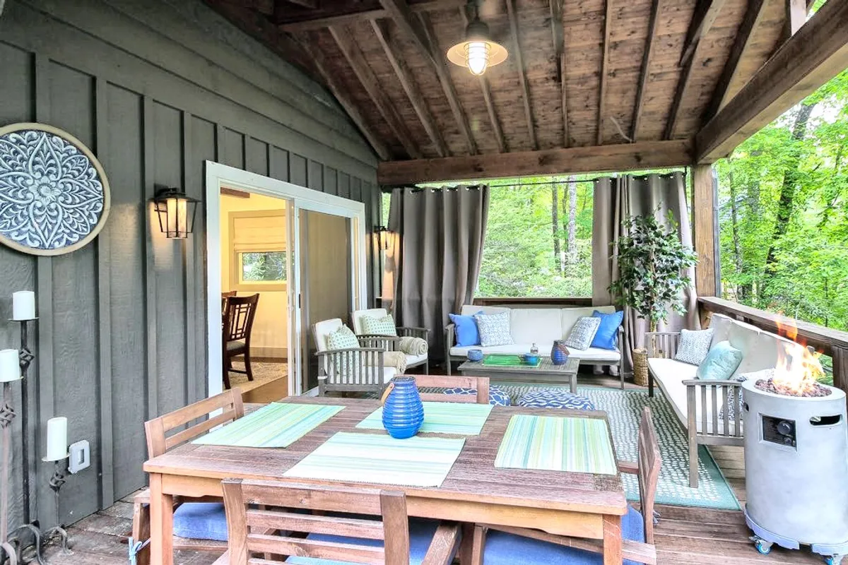 The image showcases a spacious covered deck overlooking natural scenery, with a large dining table set for gatherings, inviting outdoor seating arranged around a glowing fire pit, and tasteful decorative wall art. The warm glow of sunset filters through the trees, enhancing the tranquil and inviting atmosphere.
