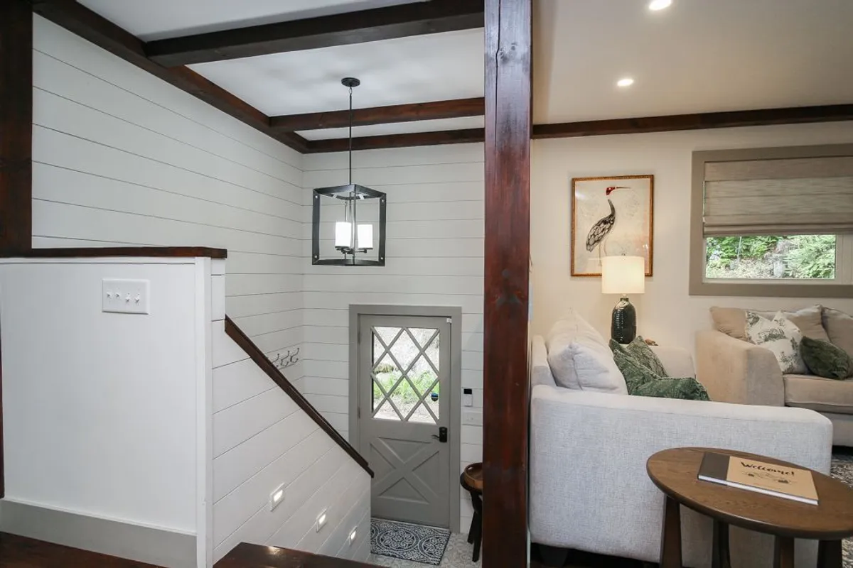 A bright and inviting interior scene focused on the staircase, front door, and a cozy seating area with artful touches. The image conveys a sense of welcome and comfort, suggesting memorable arrivals, shared moments, and the warmth of gathering with friends or a partner.
